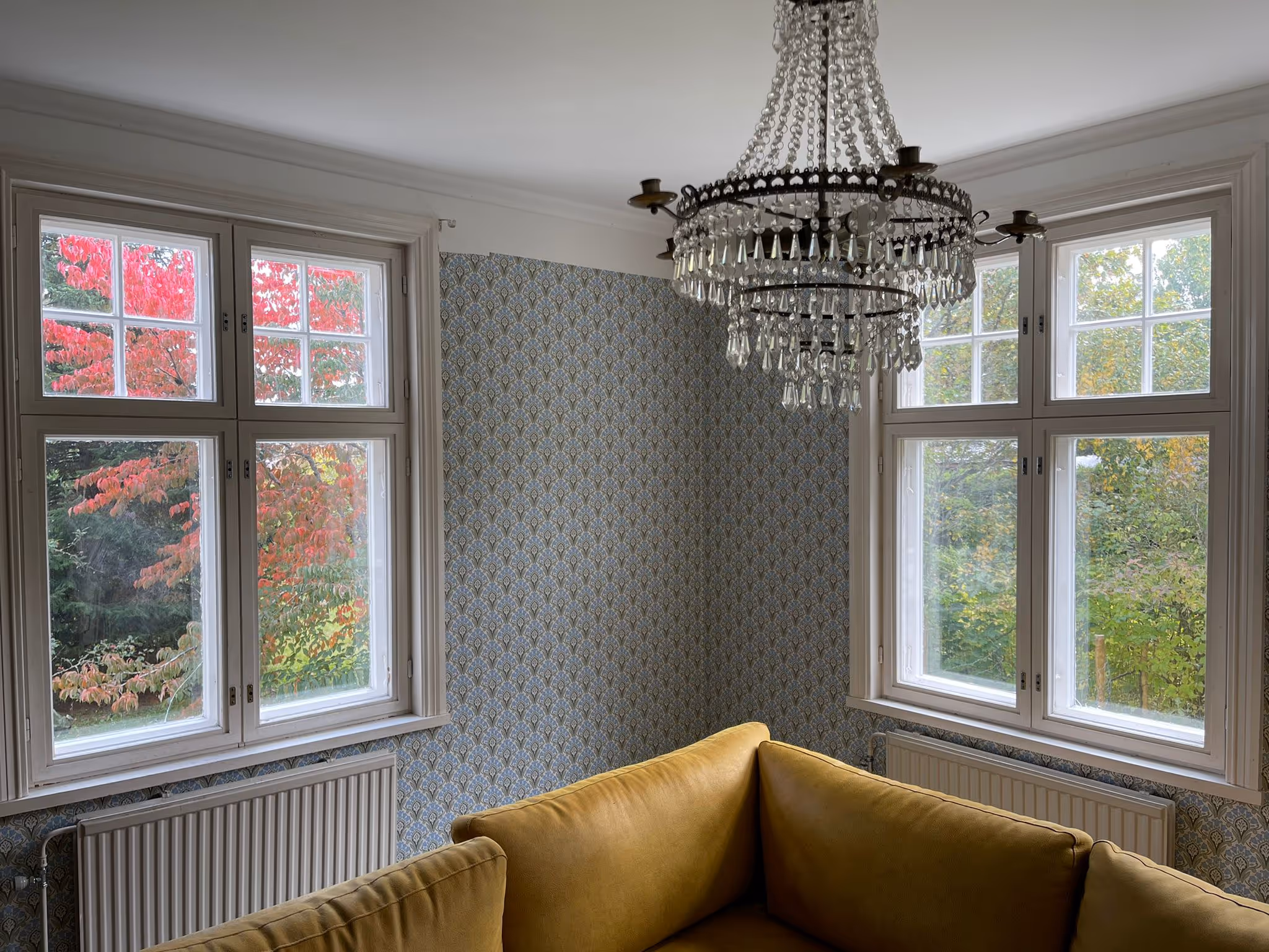 Huoneessa on kaksi ikkunaa, joista näkyy syksyinen maisema, kristallikruunu katossa ja keltainen sohva. Seinät on tapetoitu kuviollisella tapetilla.