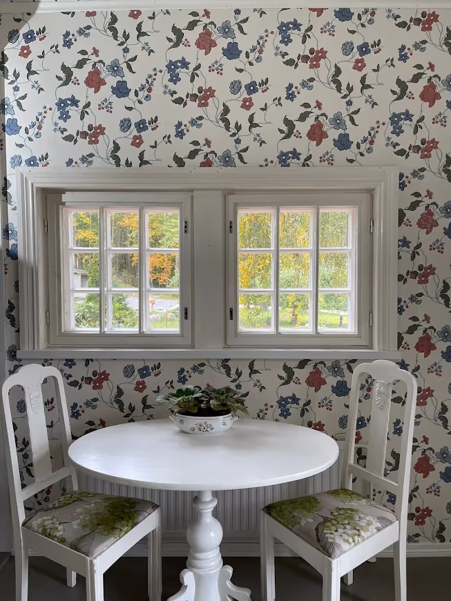 Huoneessa on kaksi valkoista tuolia, valkoinen pyöreä pöytä, jonka päällä on kukkaruukku, ja taustalla kaksi ikkunaa kukkakuvioisella tapetilla.
