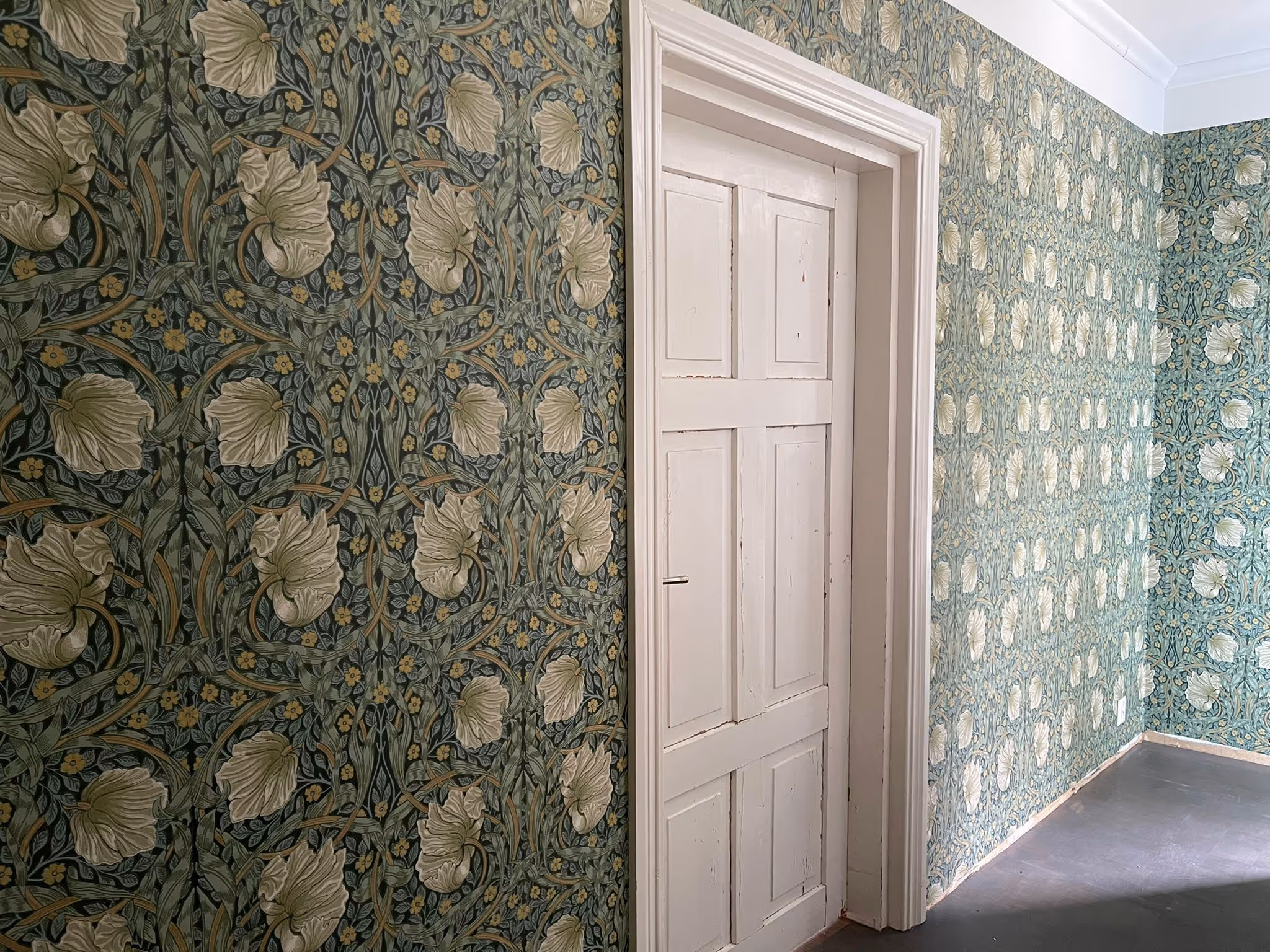 Huoneessa on kaksi seinää, jotka on tapetoitu tummanvihreällä ja vaaleankeltaisella kukkakuviolla, sekä valkoinen ovi.