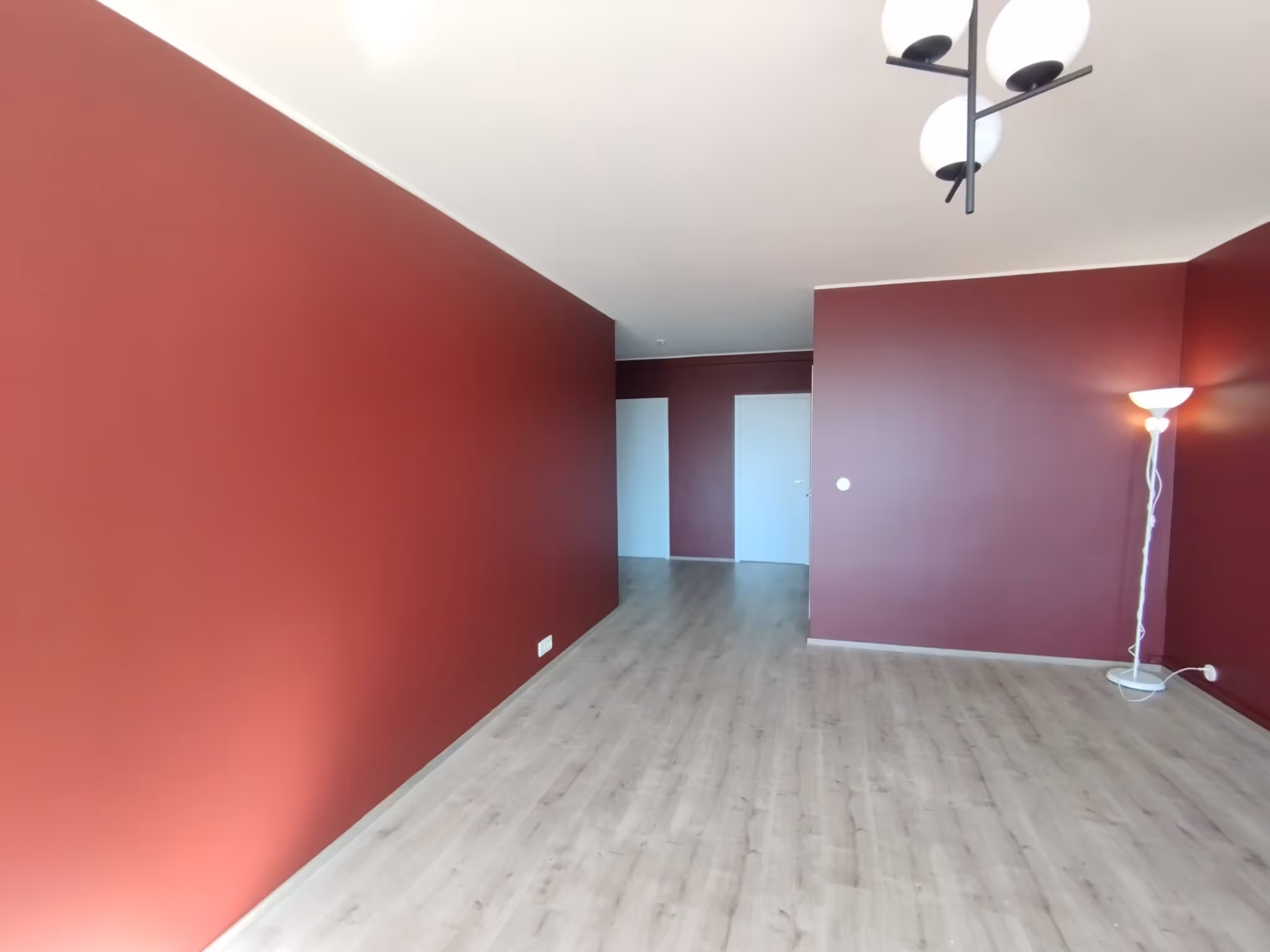 Huoneessa on punaiset seinät, vaalea laminaattilattia ja moderni kattovalaisin. Nurkassa seisoo valkoinen lattiavalaisin.