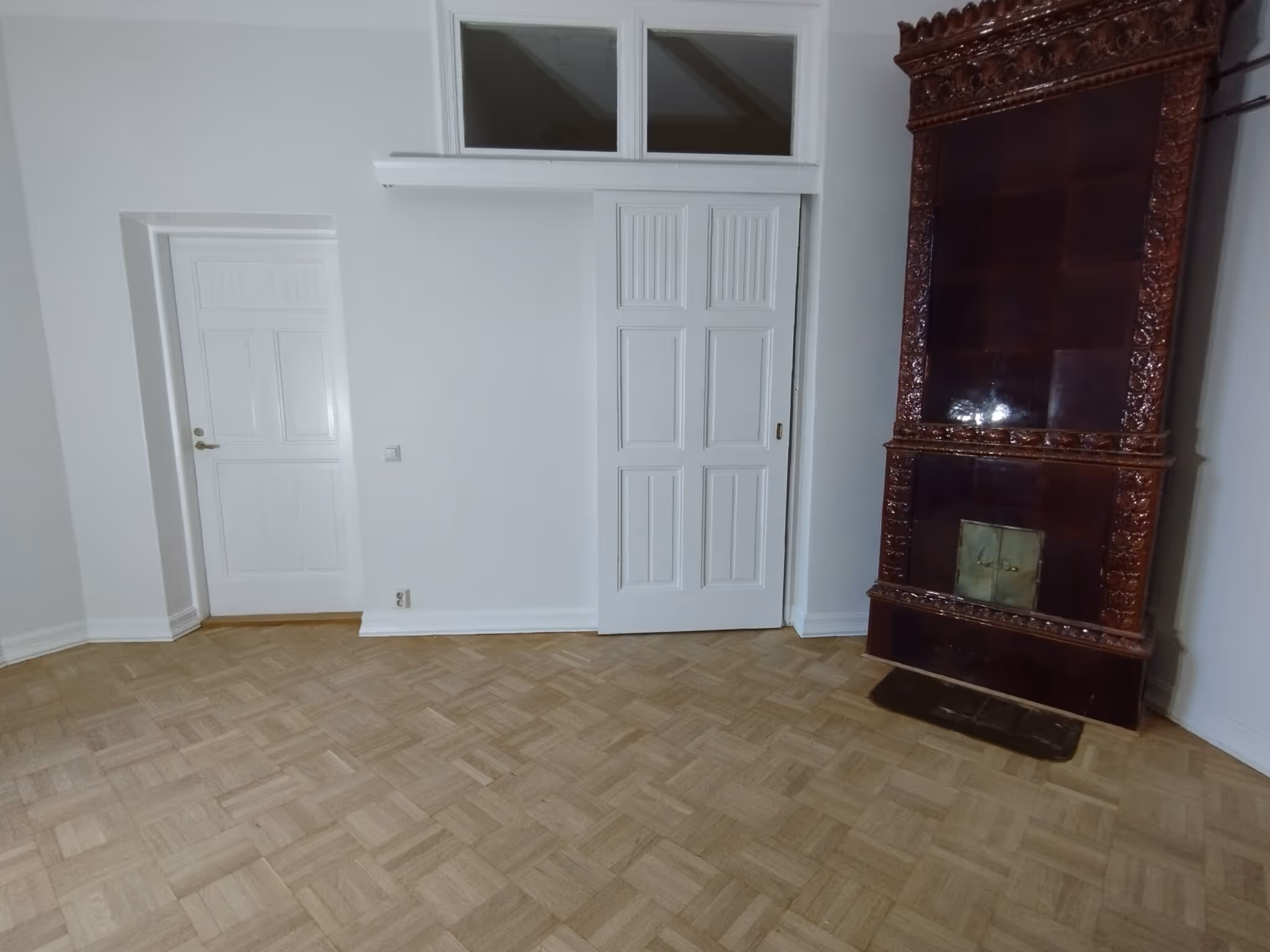 Huoneessa on vaalea parkettilattia, valkoiset seinät, kaksi valkoista ovea ja koristeellinen, tummanruskea uuni.