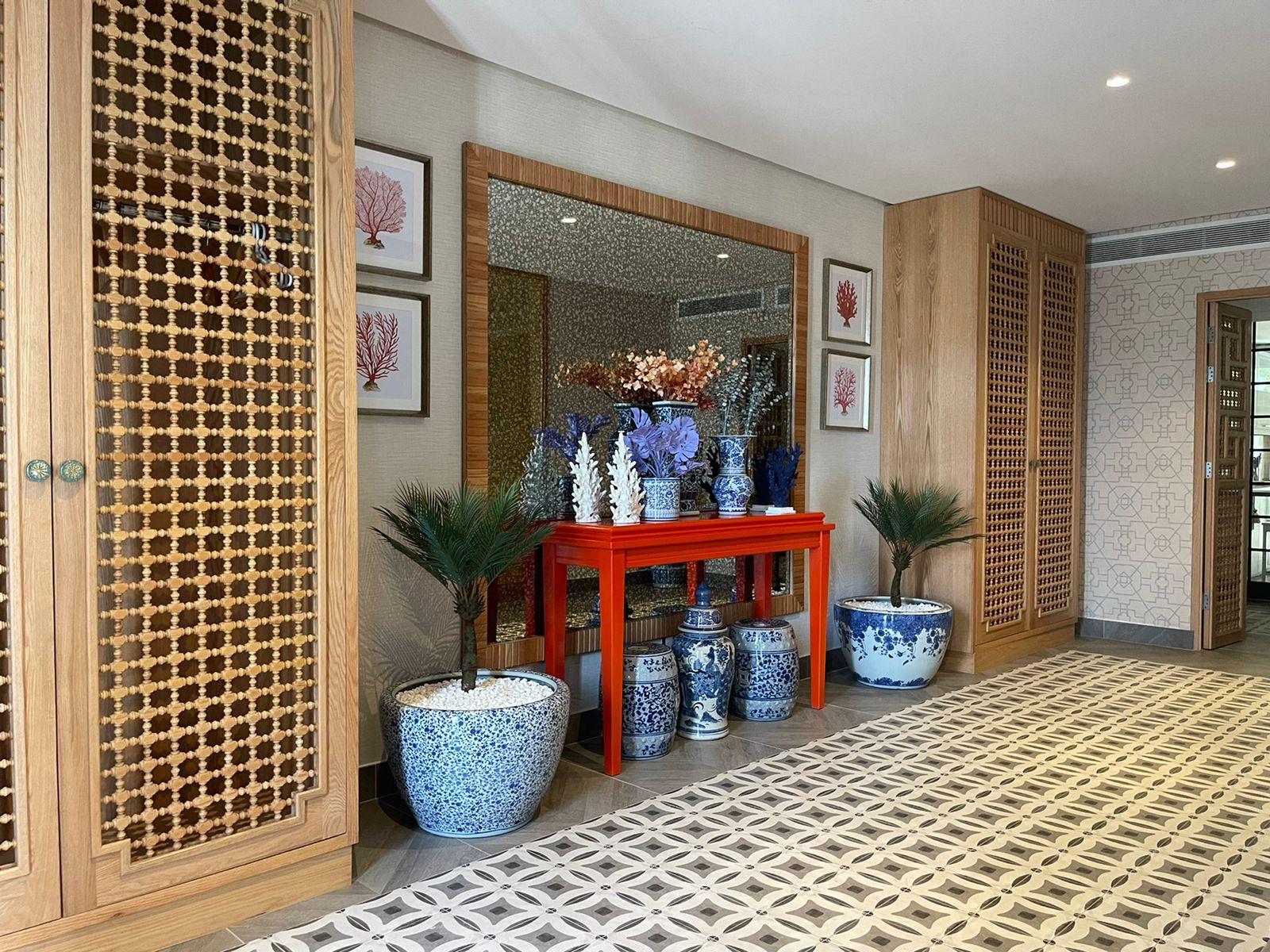 Interior hallway with patterned tile floor, wooden lattice cabinets, red table with blue and white ceramic vases, two potted plants, and framed coral artwork.
