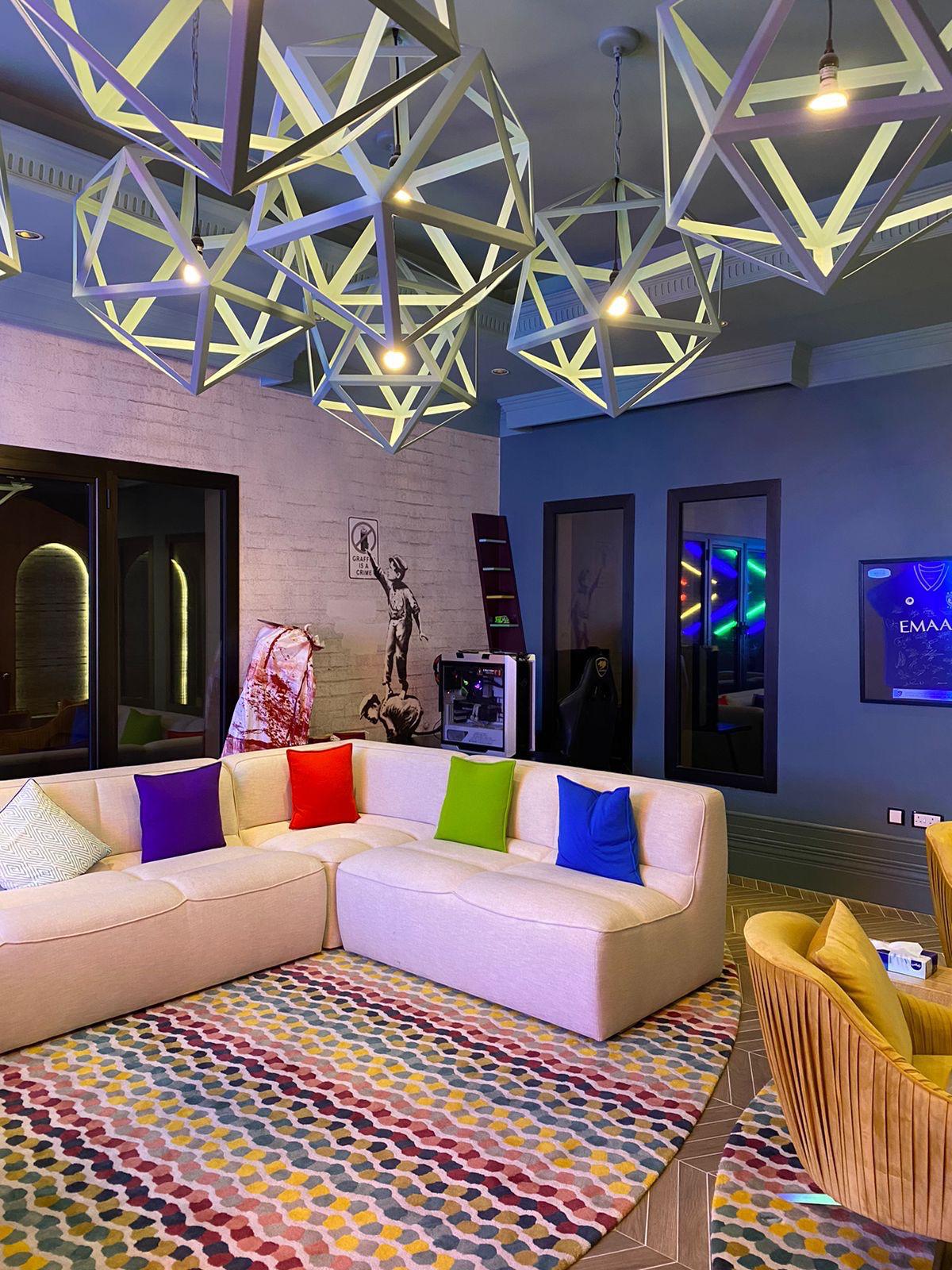 Modern living room with a beige sectional sofa featuring multicolored cushions, geometric pendant lights, and a colorful patterned rug.