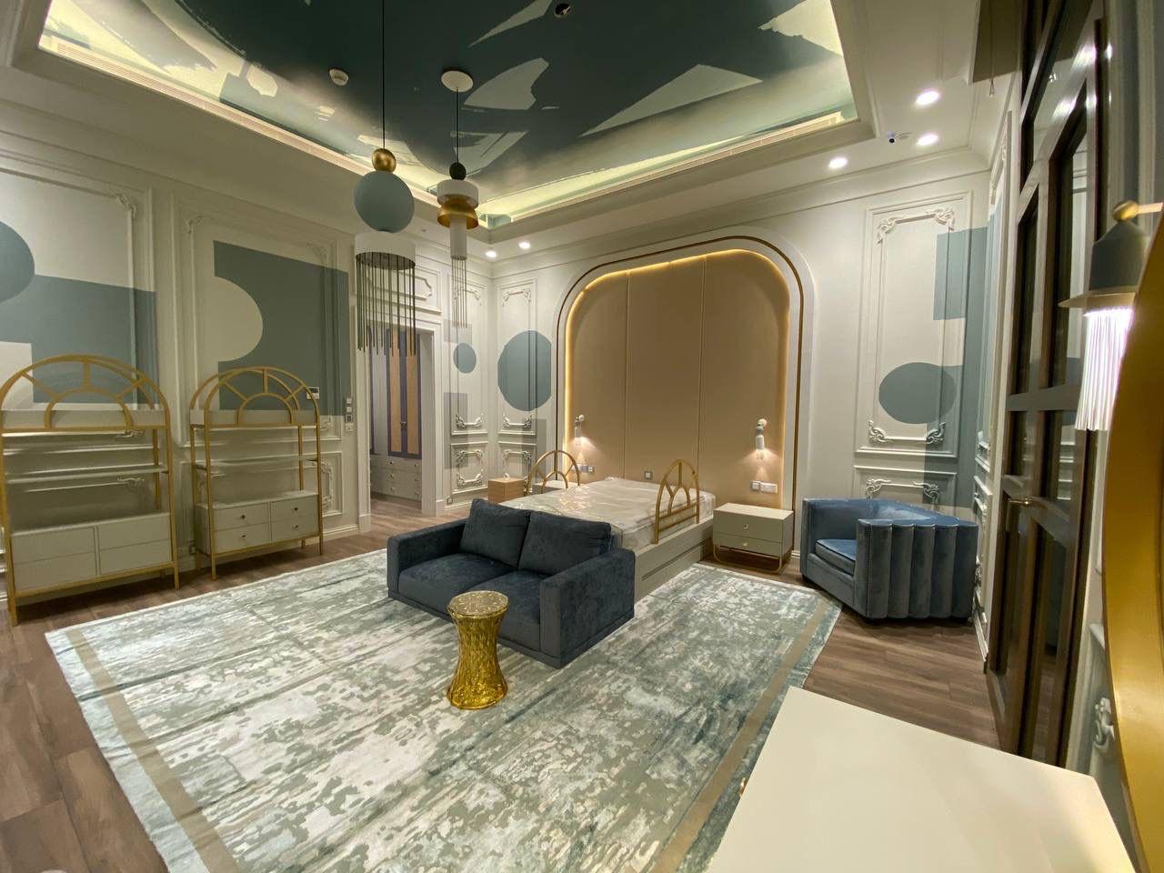 Modern bedroom with double bed, blue sofa, gold side table, blue armchair, patterned rug, and soft wall lighting.