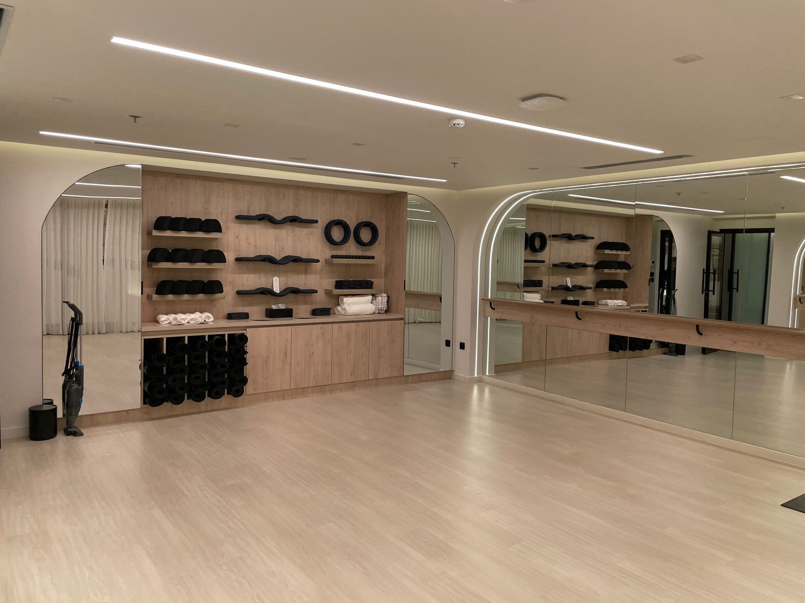 Modern fitness studio with wooden storage shelves holding black yoga mats, workout rollers, and white towels, a large mirror wall with ballet barre, and light wood flooring.