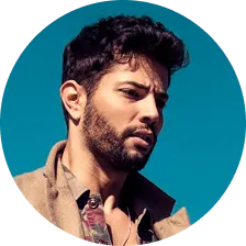 Close-up of a young man with dark hair and beard against a clear blue sky.