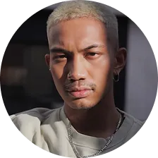 Portrait of a young man with short bleached blonde hair, wearing a silver chain necklace and an earring, looking directly at the camera.