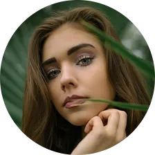 Close-up portrait of a young woman with long brown hair looking at the camera behind green leaves.