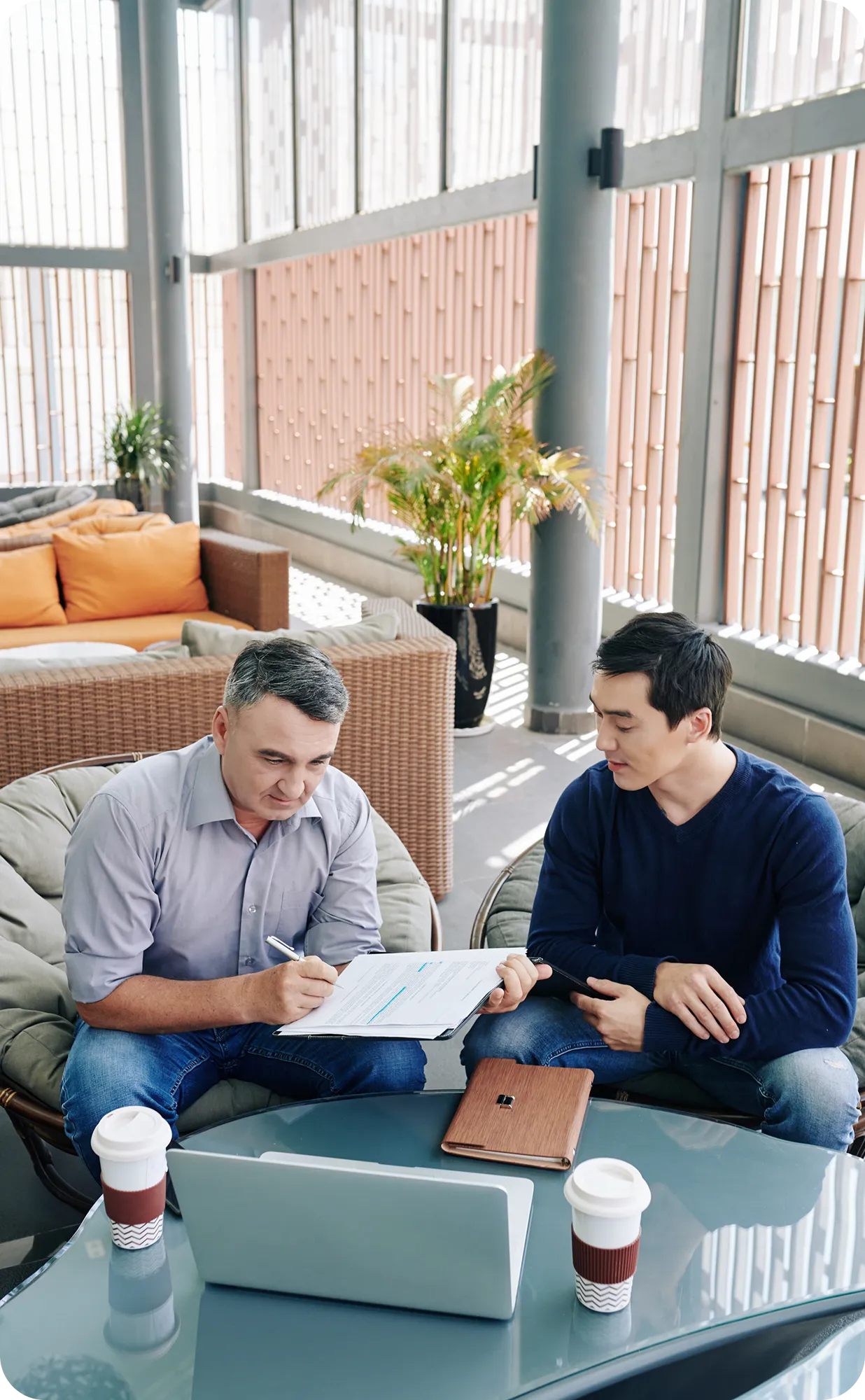 Two men sitting in chairs at a glass table with coffee cups and a laptop, reviewing documents together in a bright indoor space.