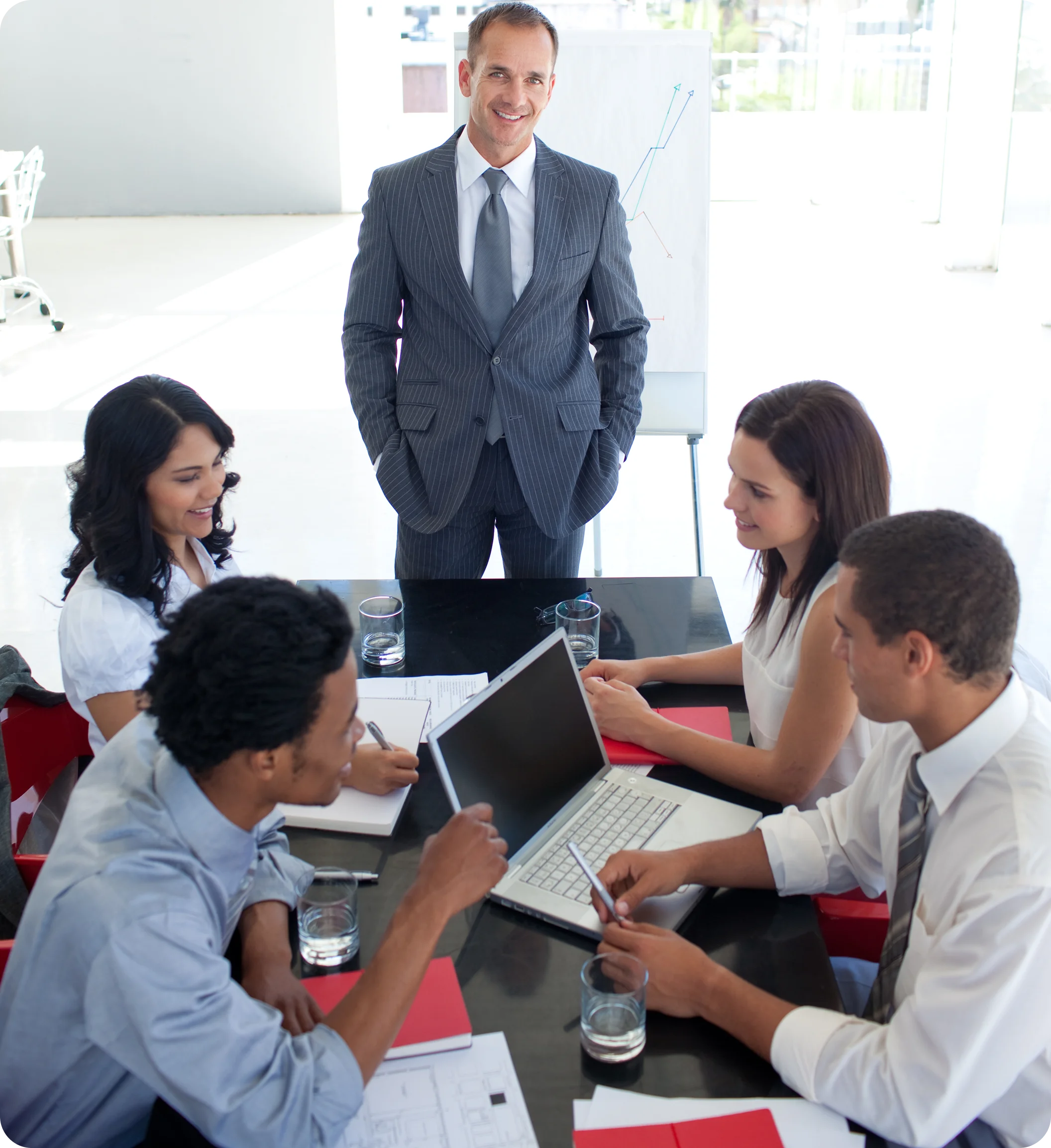 Businessman standing and smiling while four colleagues sit around a table with a laptop, notebooks, and glasses of water in a bright office.