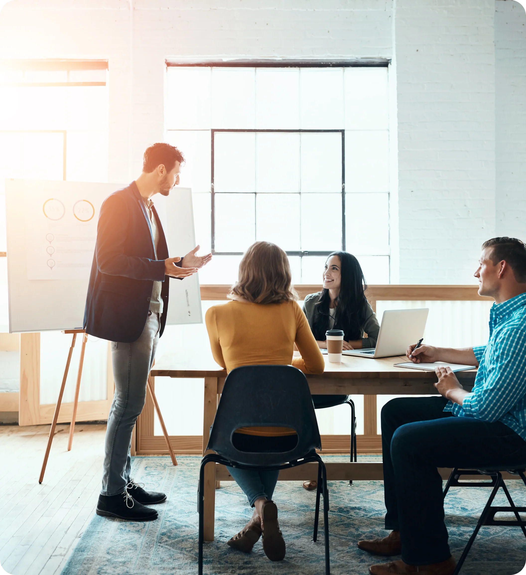 A man stands presenting to three colleagues seated at a table with a laptop and coffee cup in a bright office.