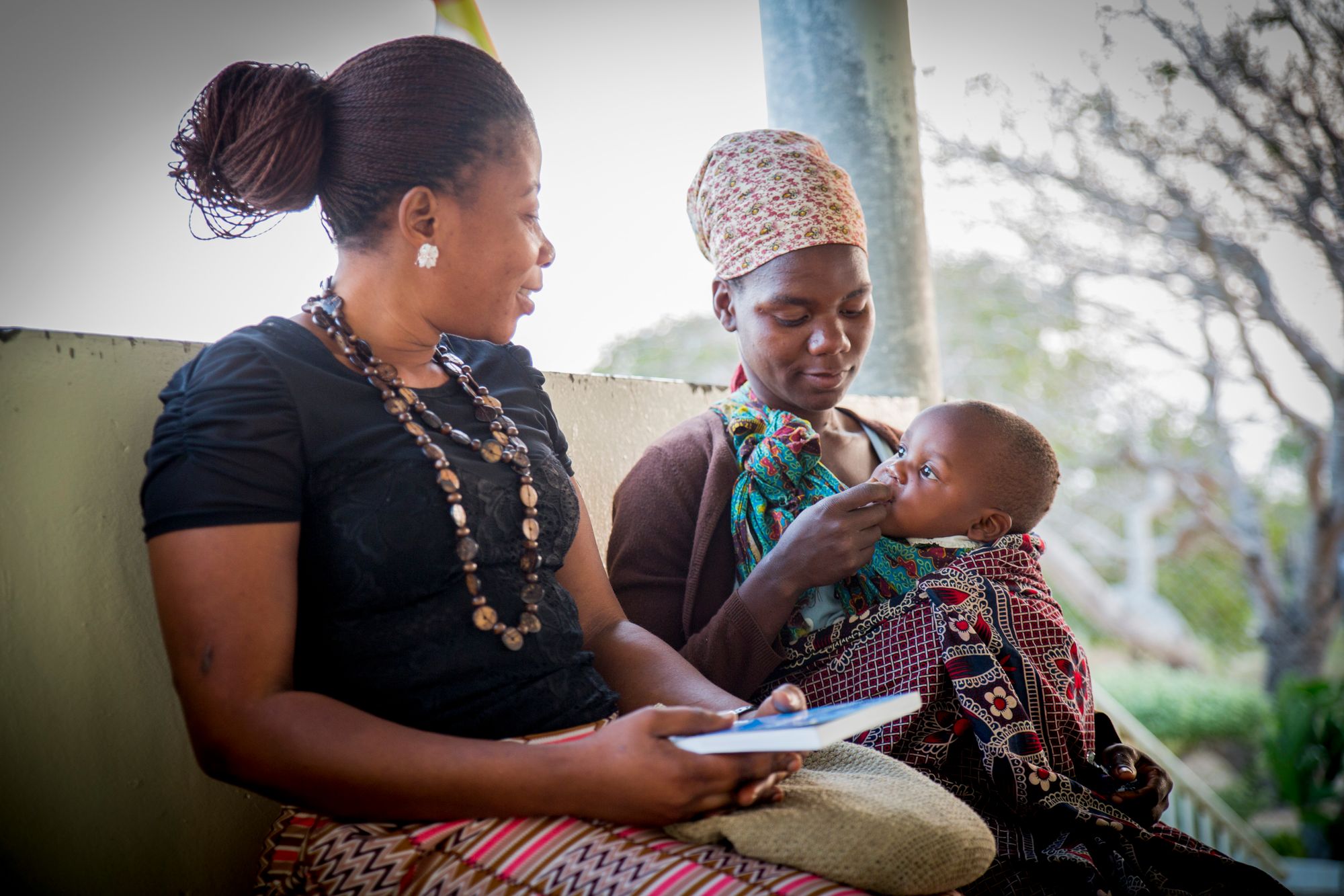 A health advocate with a notebook sits beside a mother feeding her baby on a health centre veranda