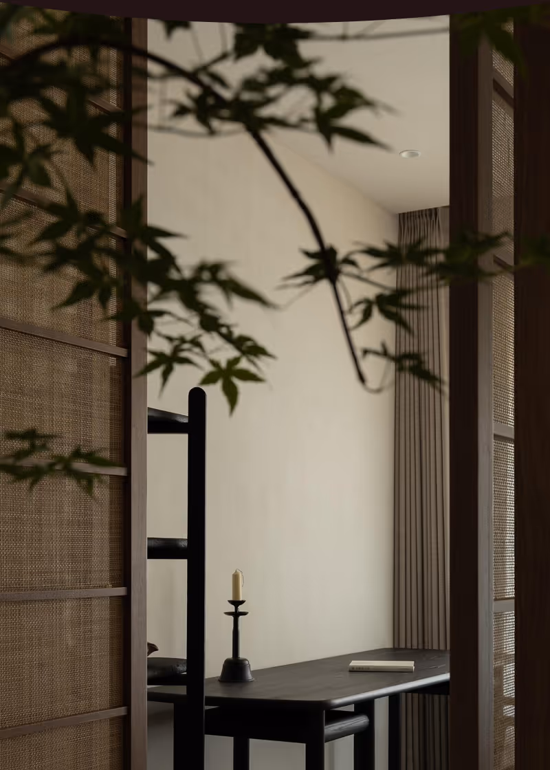 Minimalist desk with a black candle holder and a closed book, viewed through branches and framed by wooden mesh panels.