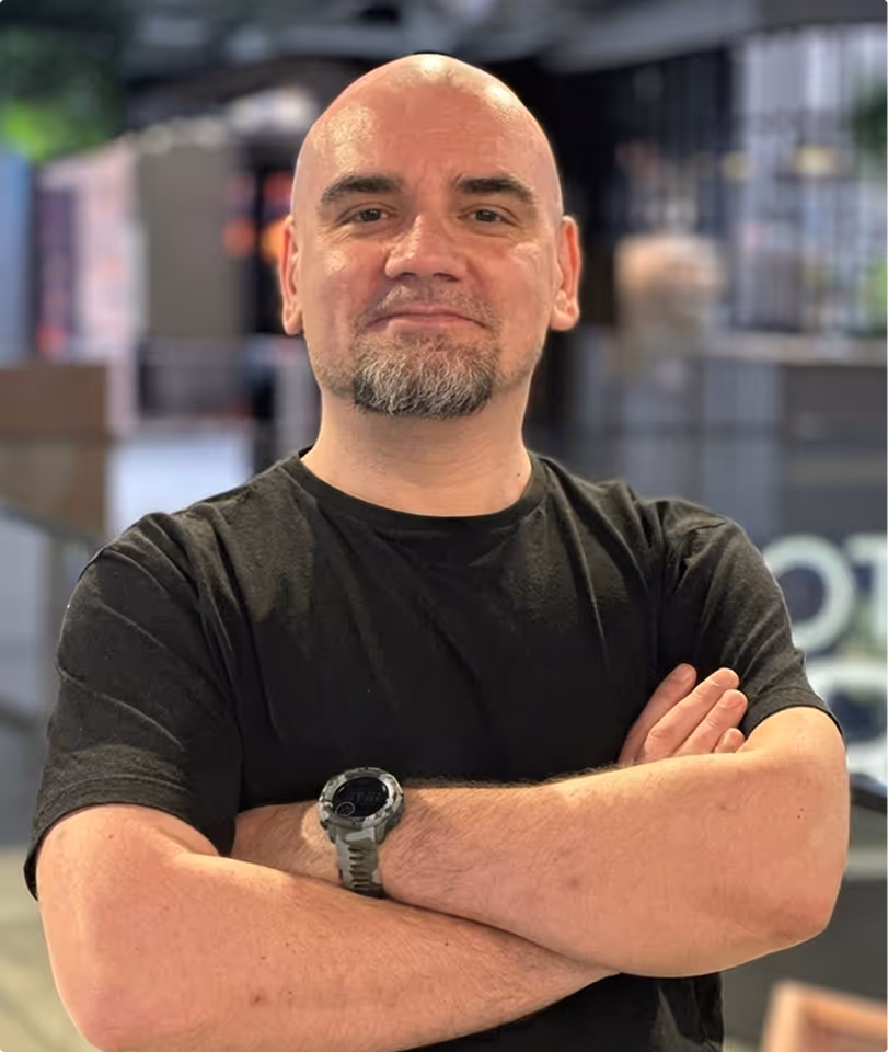 Bald man with a goatee wearing a black t-shirt and a smartwatch, standing with arms crossed in a blurred indoor setting.