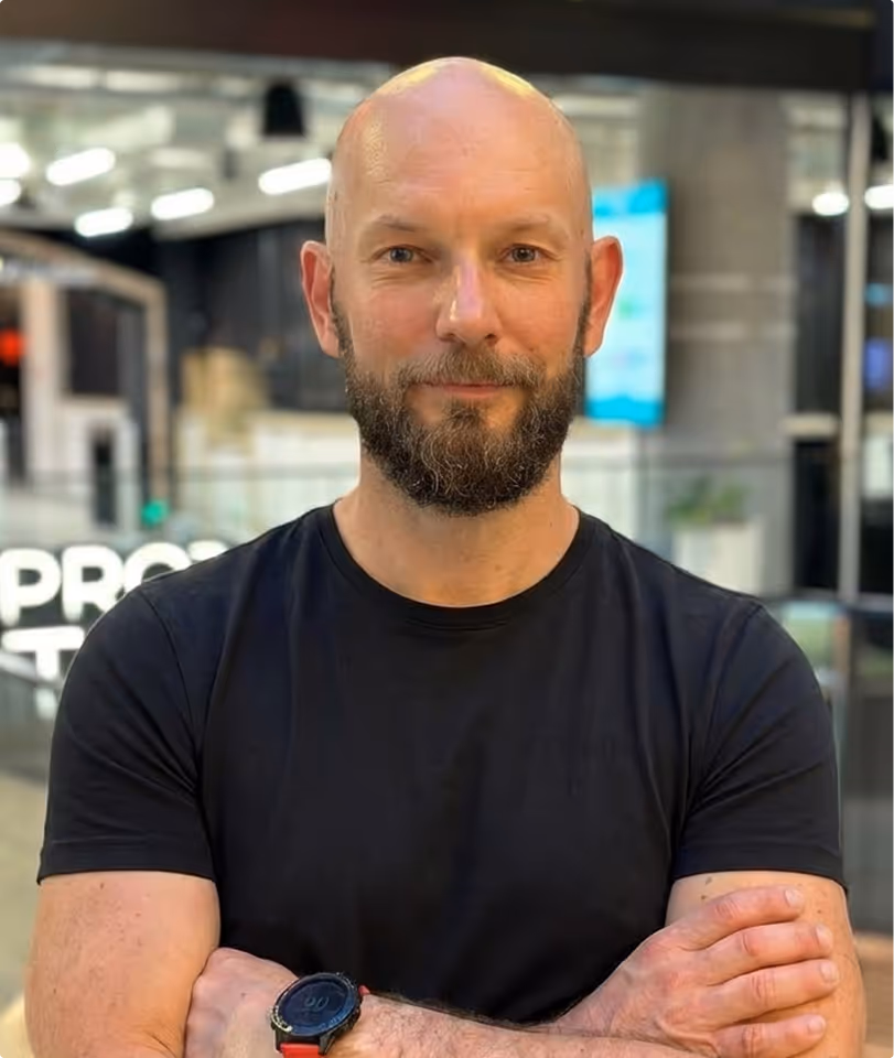 Bald man with a beard wearing a black t-shirt and a wristwatch, standing with arms crossed indoors.