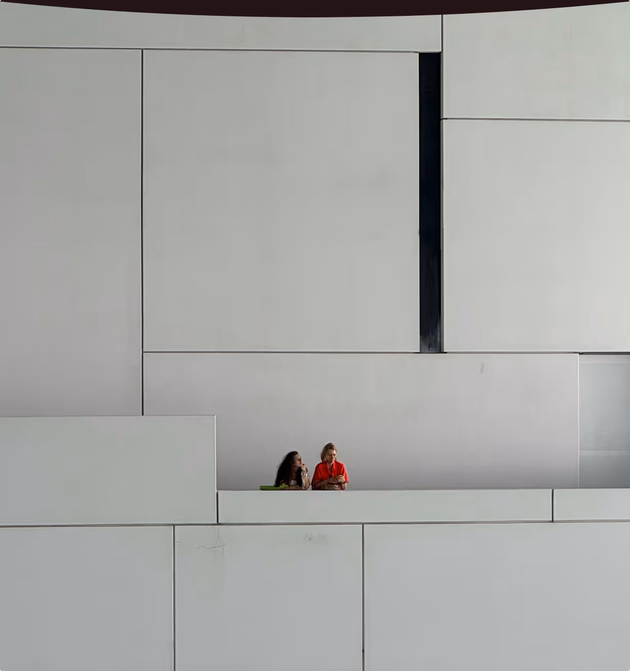 Two women sitting and looking at a phone against a large, geometric, minimalist concrete wall.