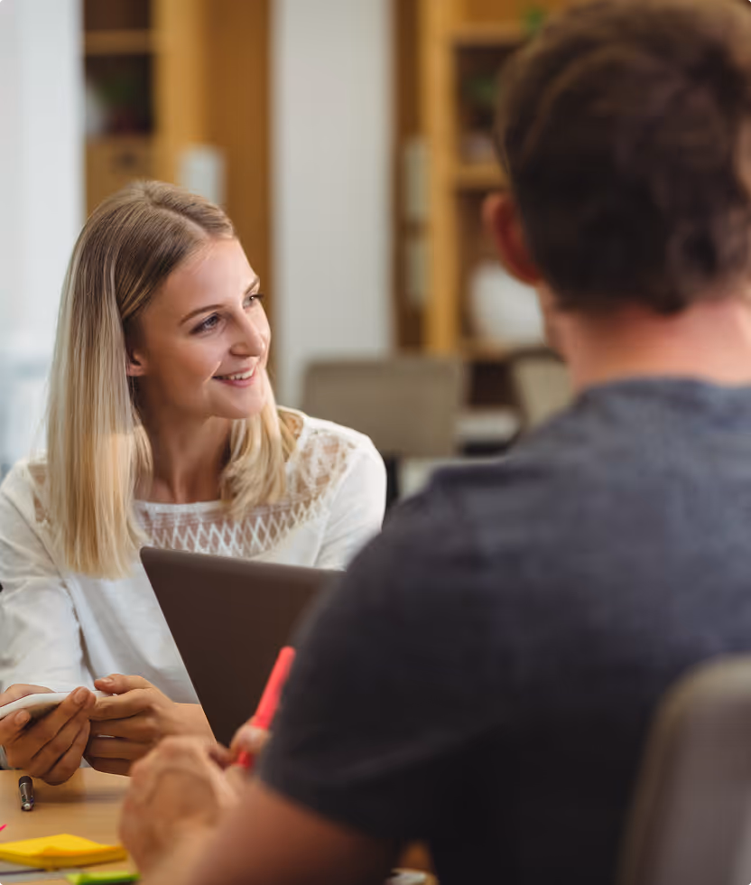 Smiling woman holding a smartphone sitting across a man with a laptop and red pen at a desk in an office.