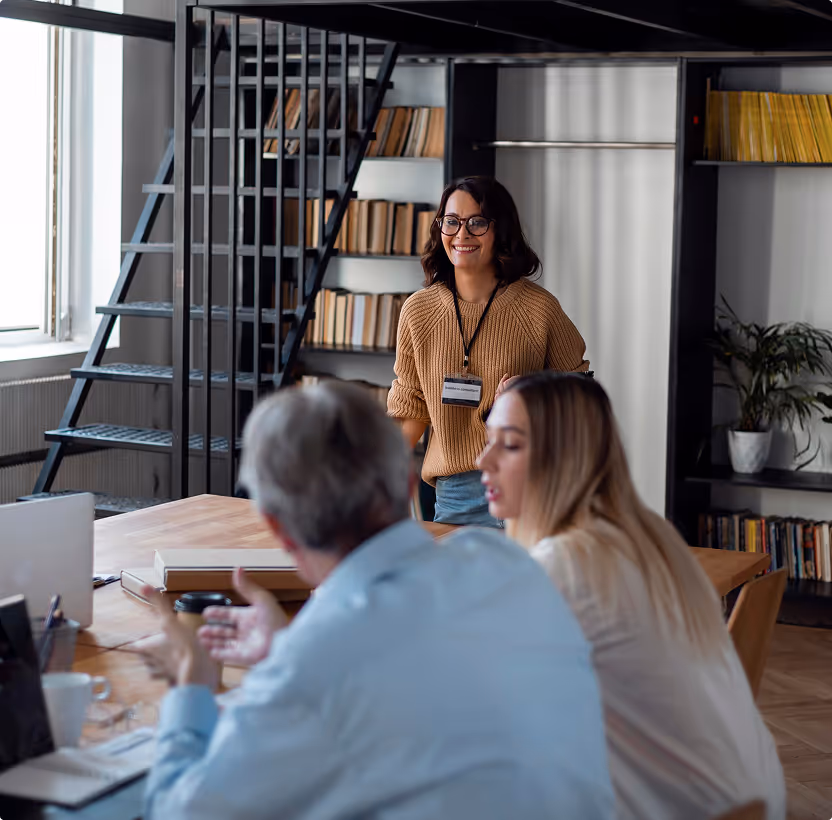 Smiling woman wearing glasses and an ID badge standing near a bookshelf while two colleagues discuss at a table.