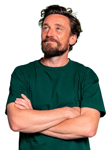 Hombre con barba y cabello rizado vestido con camiseta verde, con los brazos cruzados mirando hacia arriba y sonriendo ligeramente.