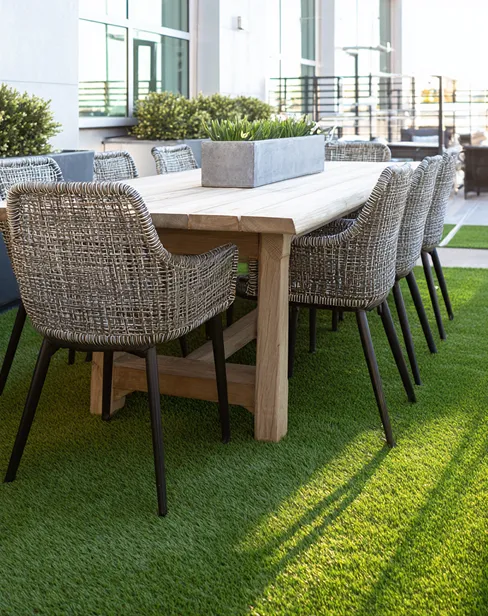 Mesa de madera clara con ocho sillas de mimbre gris en un suelo de césped artificial.