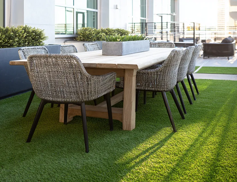 Mesa de comedor de madera con ocho sillas de mimbre gris sobre césped artificial en una terraza moderna.
