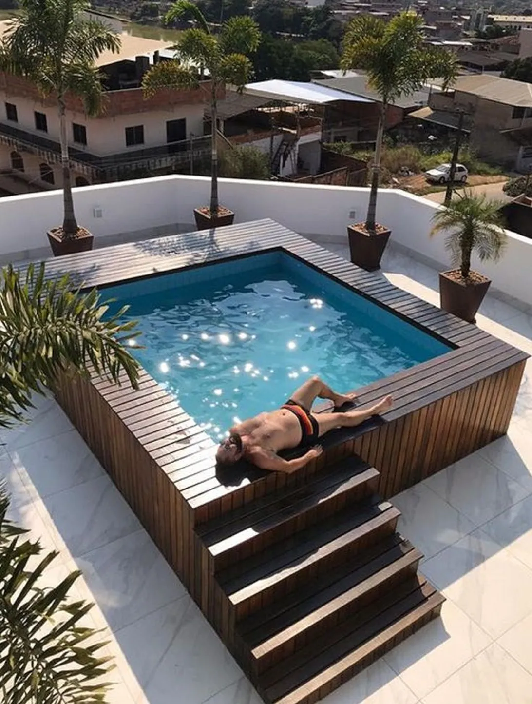 Hombre recostado junto a una piscina elevada de madera, rodeada de cuatro palmeras en macetas.