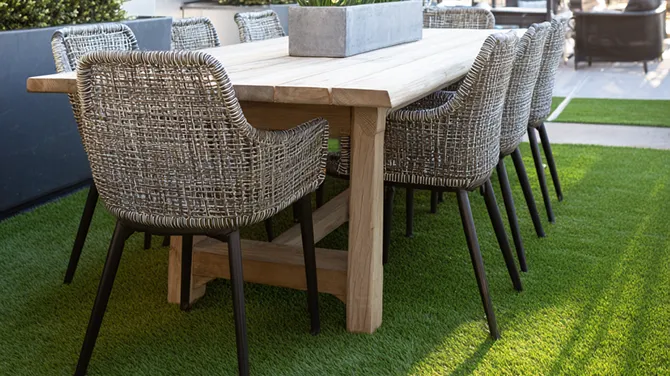Mesa de comedor de madera clara con ocho sillas de mimbre y patas negras sobre césped artificial.