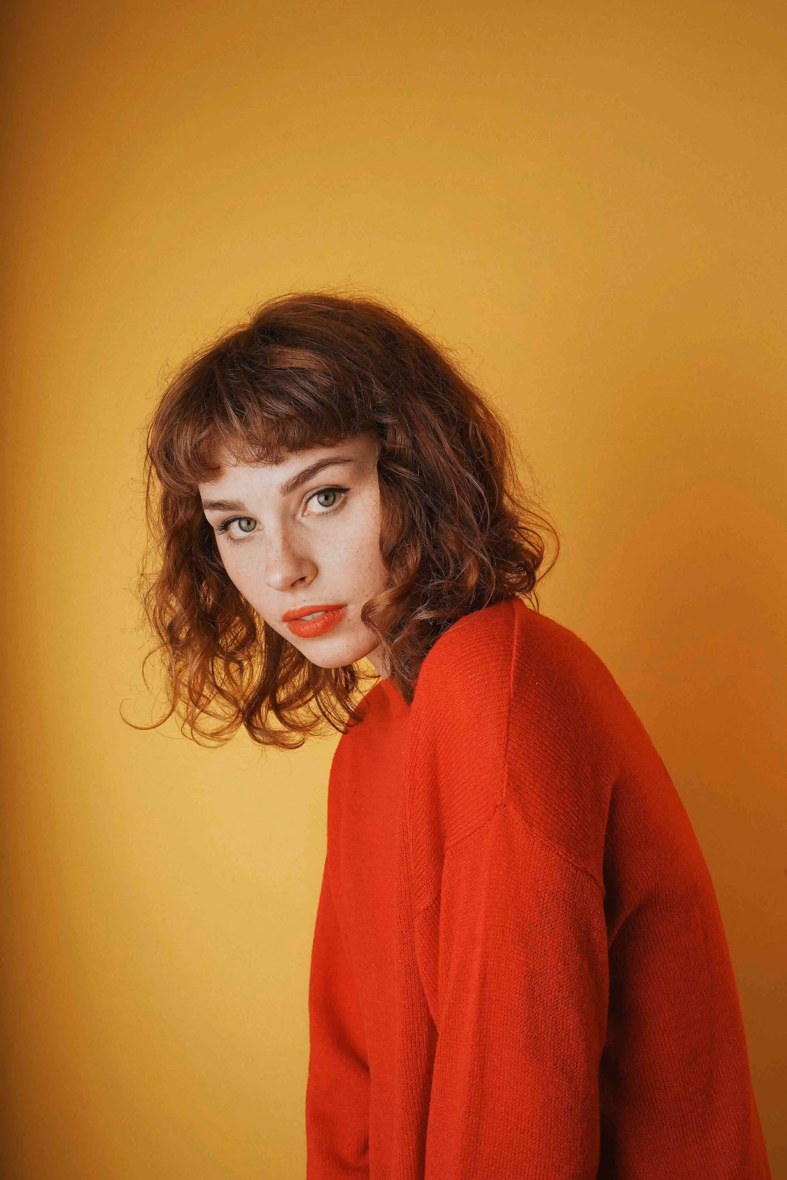 Frau mit roten, welligen Haaren und rotem Pullover vor einem gelben Hintergrund.