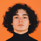 Person with curly dark hair wearing a black turtleneck against an orange background.