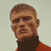 Close-up portrait of a man with short blond hair wearing a dark turtleneck and red jacket against a muted background.