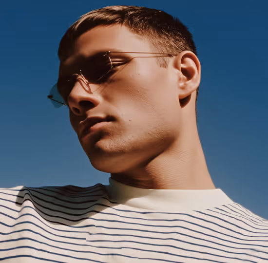 Close-up of a young man wearing thin metal-framed glasses and a white striped shirt against a clear blue sky.
