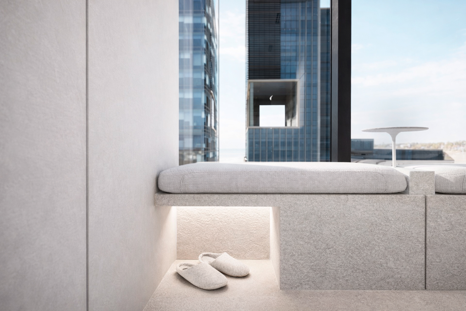Modern minimalist seating area with a cushion and a pair of slippers beneath, next to a large window overlooking tall buildings.
