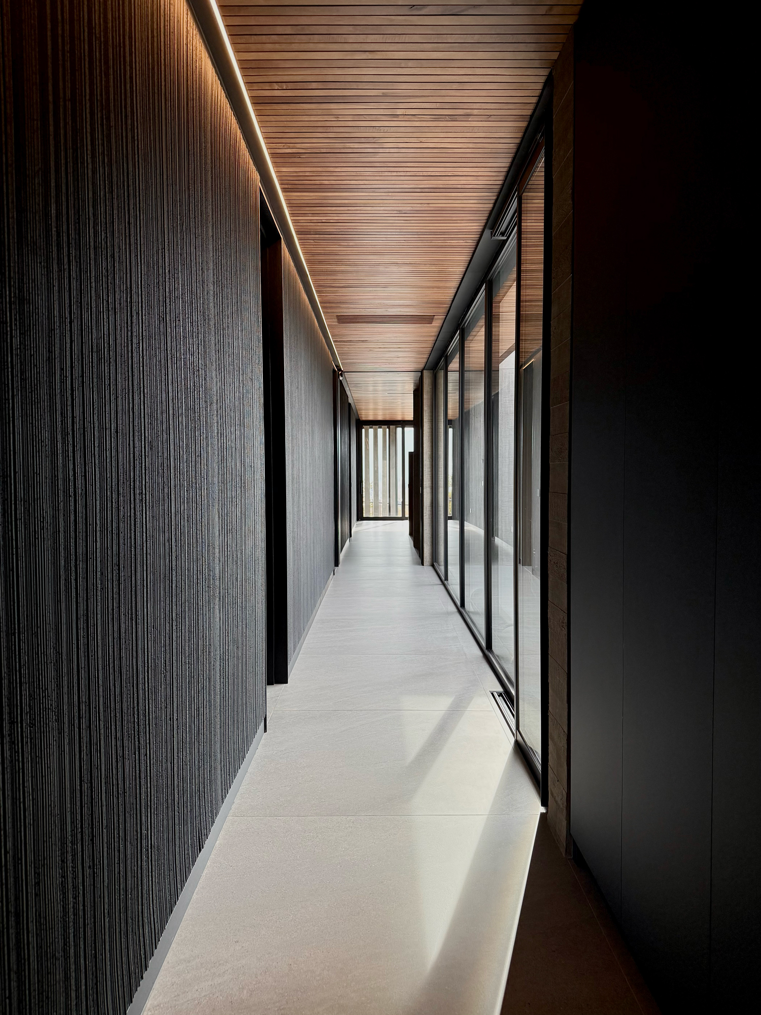 Modern hallway with textured dark wall on the left, large glass windows on the right, and wooden slatted ceiling.