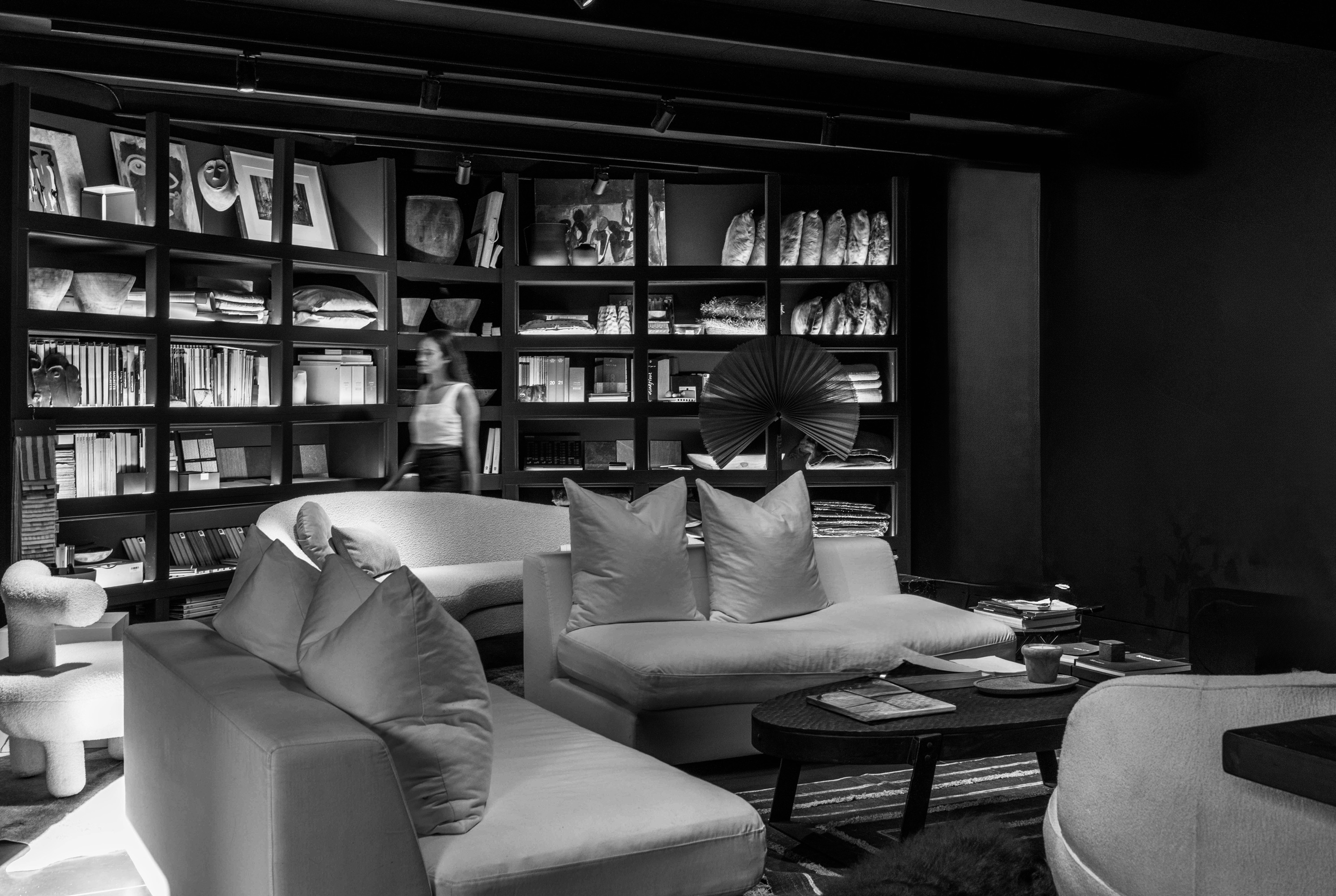 Modern living room with plush sofas, a coffee table, and a wall of shelves filled with books, decor items, and a blurred woman walking.