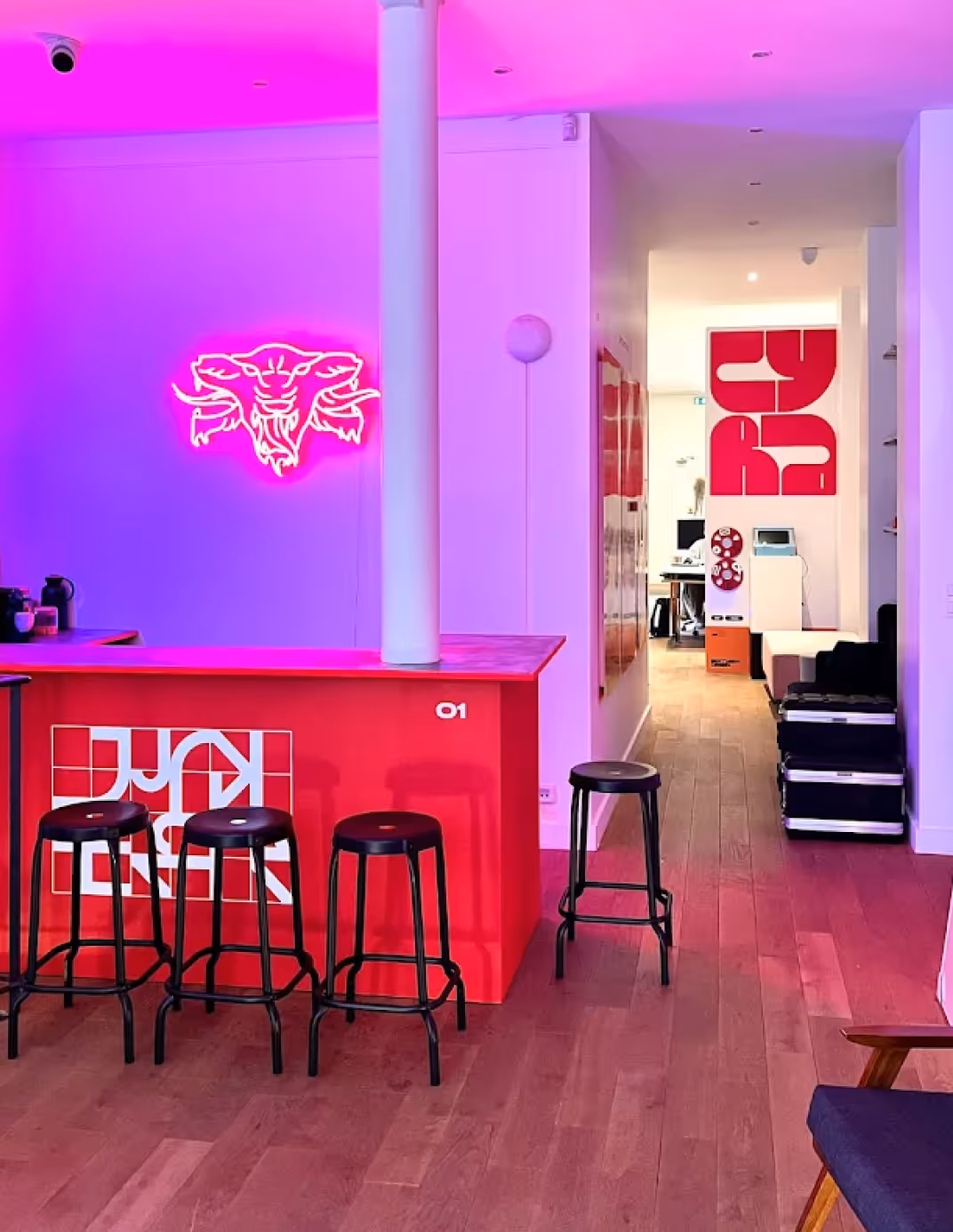 Intérieur moderne avec un bar rouge éclairé par une lumière rose, quatre tabourets noirs, un mur avec un néon en forme de tête de taureau, et un espace de bureau à l'arrière avec un grand panneau mural rouge.