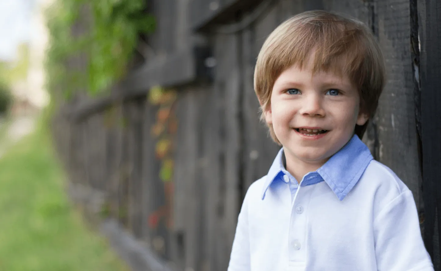 A young child stands beside a black wooden fence, wearing a white shirt with a blue collar, surrounded by greenery.