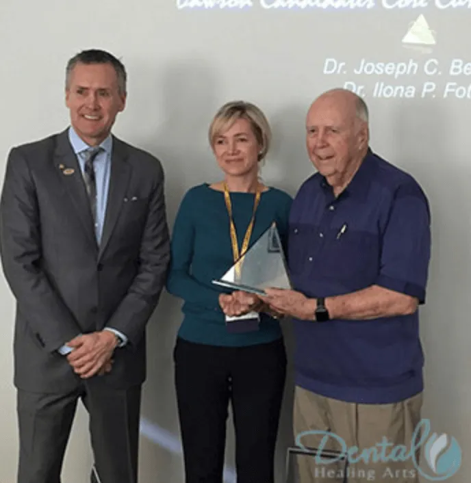 Three professionals stand together, holding an award trophy, with a background displaying text related to dental arts.