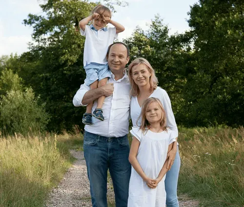 A family stands together on a grassy path, with a child playfully perched on a parent's shoulders, enjoying a sunny day outdoors.