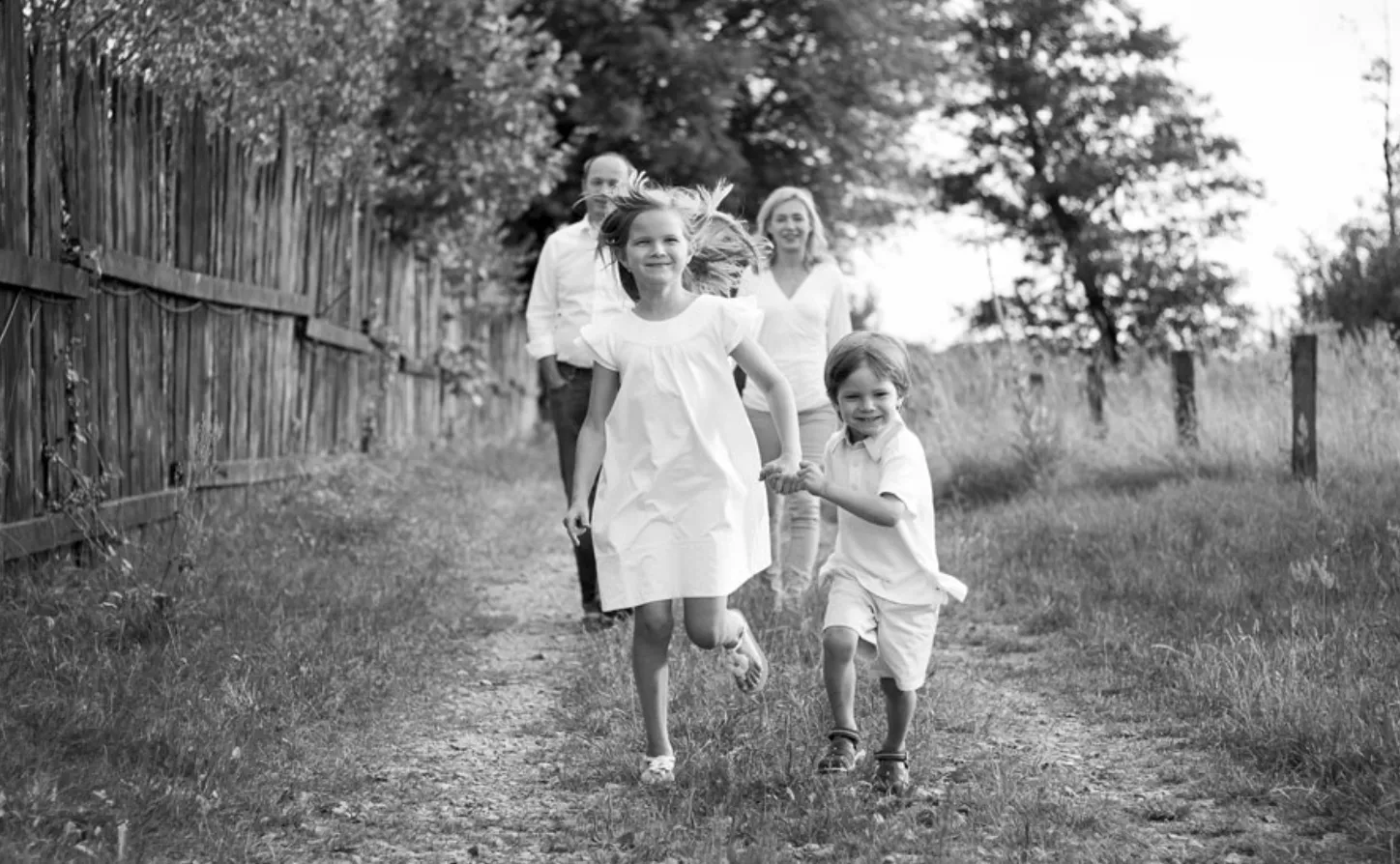 A group of children joyfully run down a gravel path surrounded by greenery and a wooden fence, capturing a moment of carefree adventure.