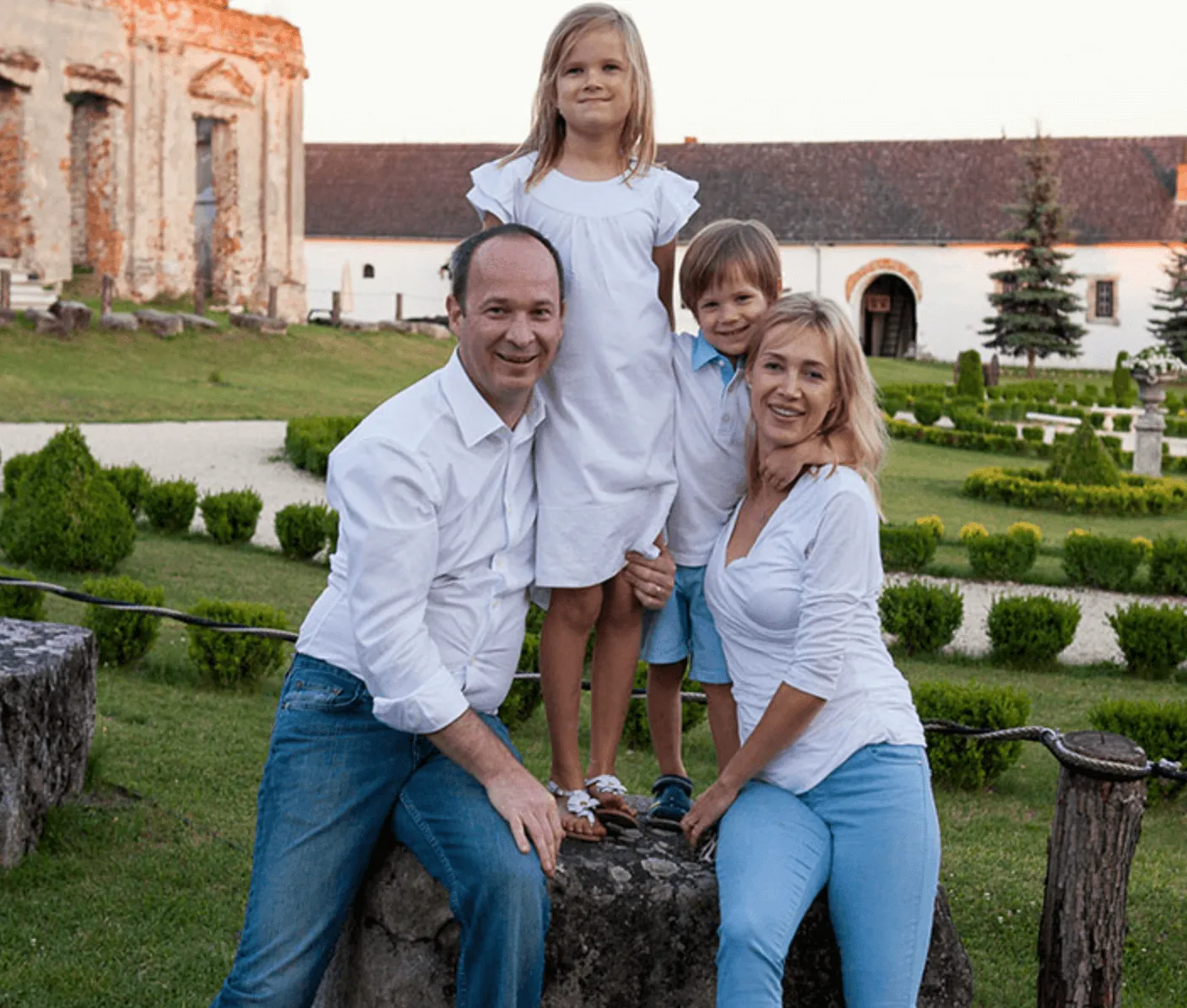 A family of four dresses in white poses outdoors near a historic building, surrounded by meticulously landscaped greenery.