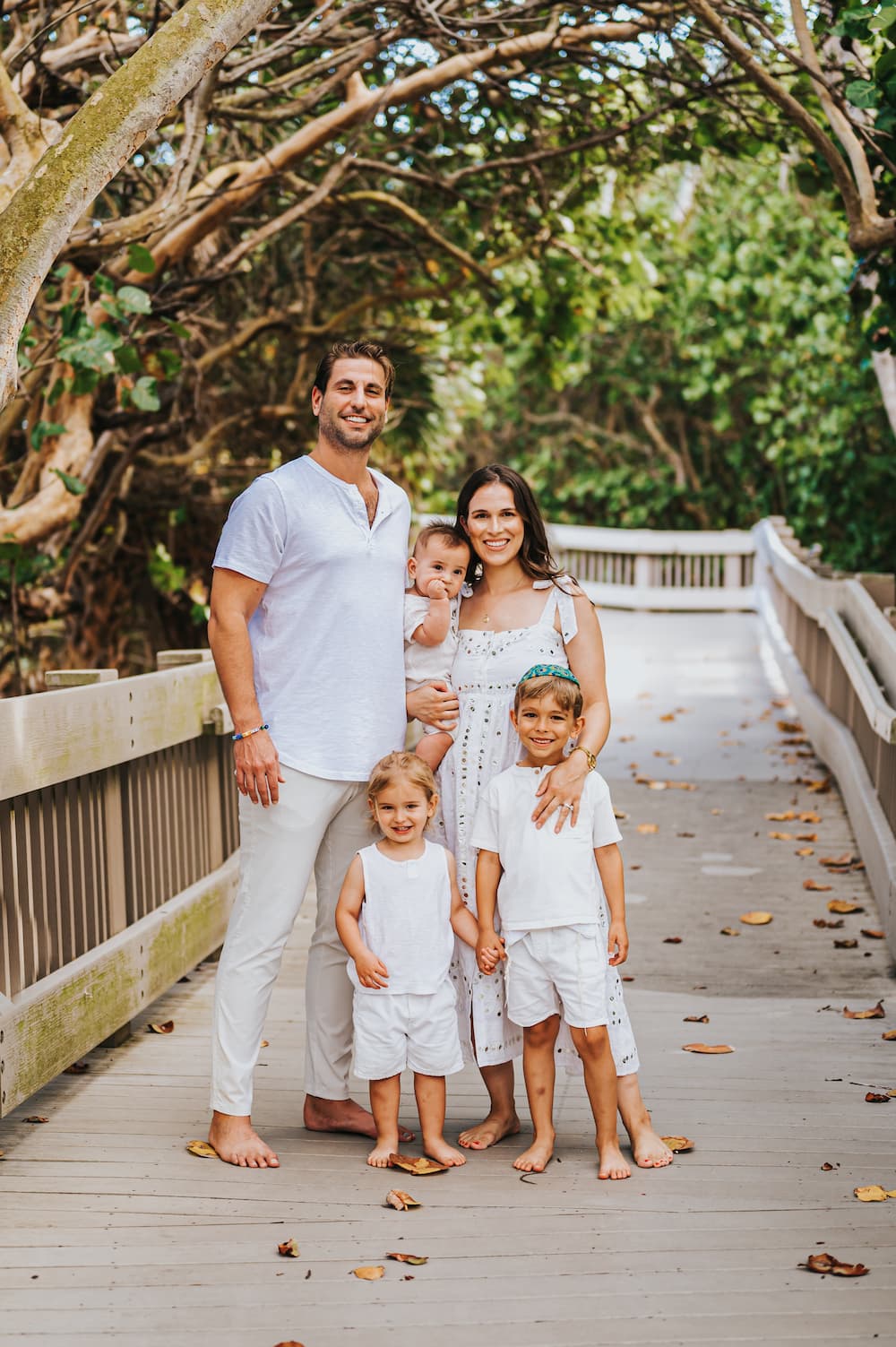 A family of four stands on a wooden walkway surrounded by lush green foliage, wearing coordinated white outfits.