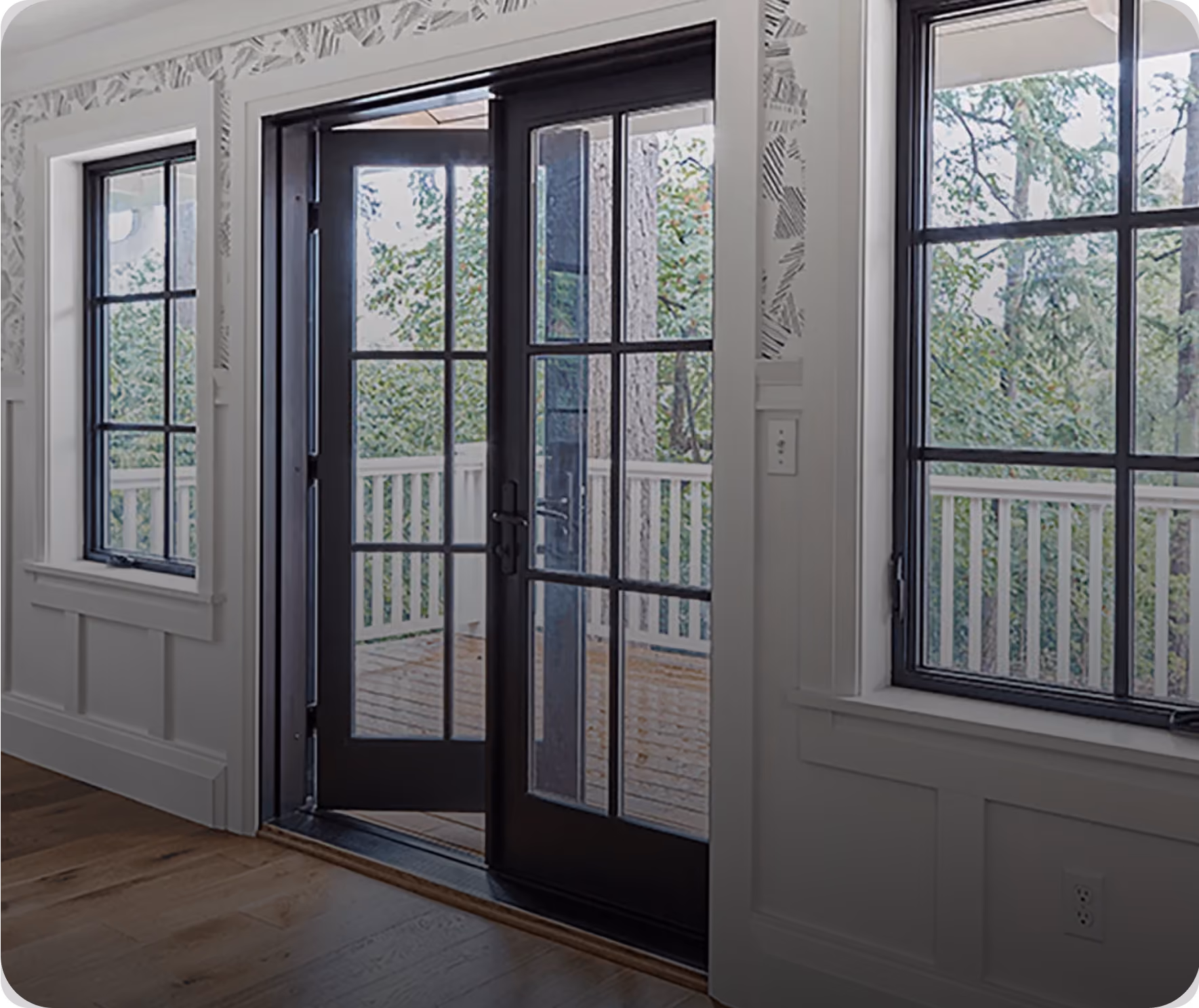 Interior view of a white room featuring two black-framed windows and a set of black-framed French doors opening to a wooden deck surrounded by green trees.