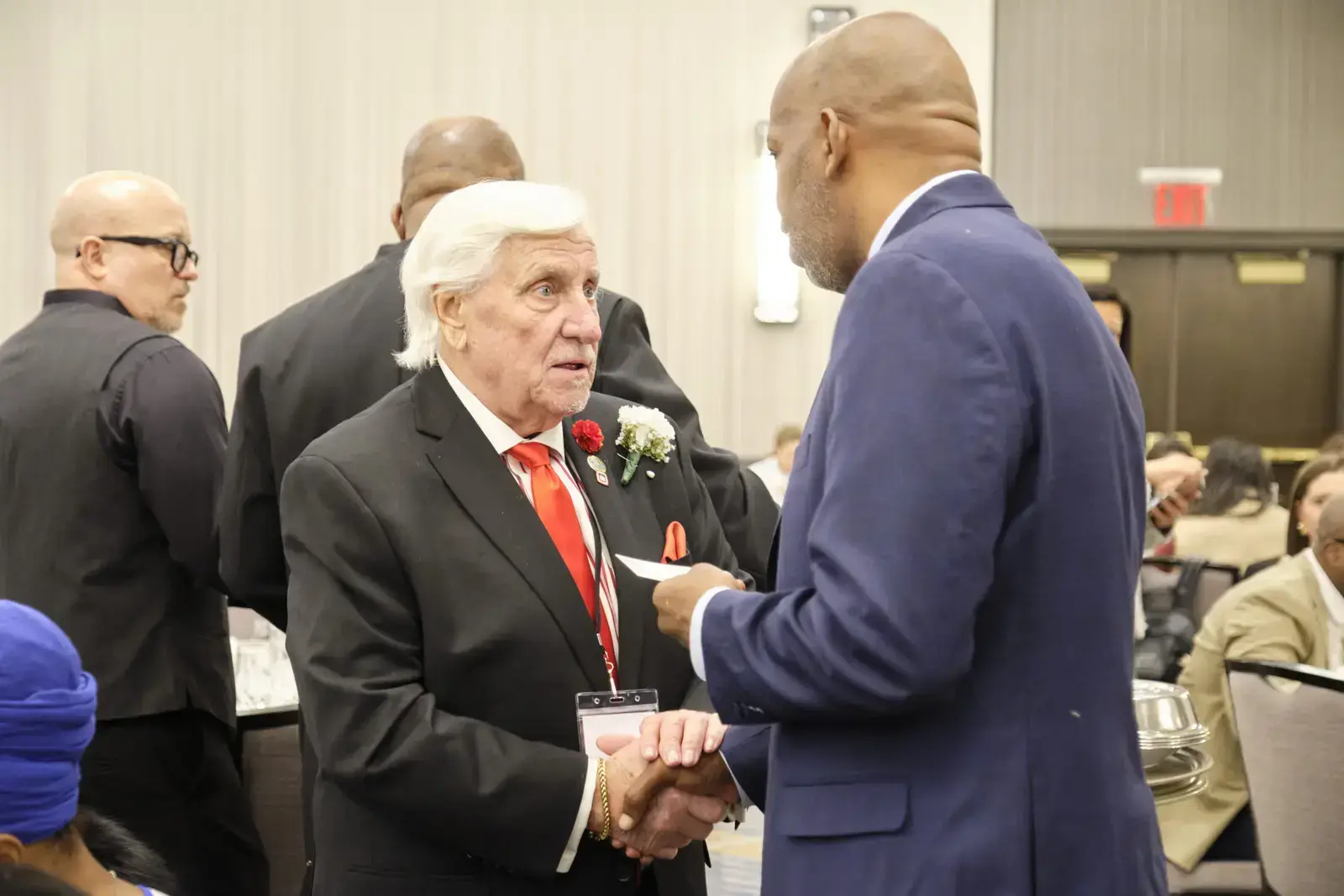 Older man with corsage shaking hands with attendee at formal event