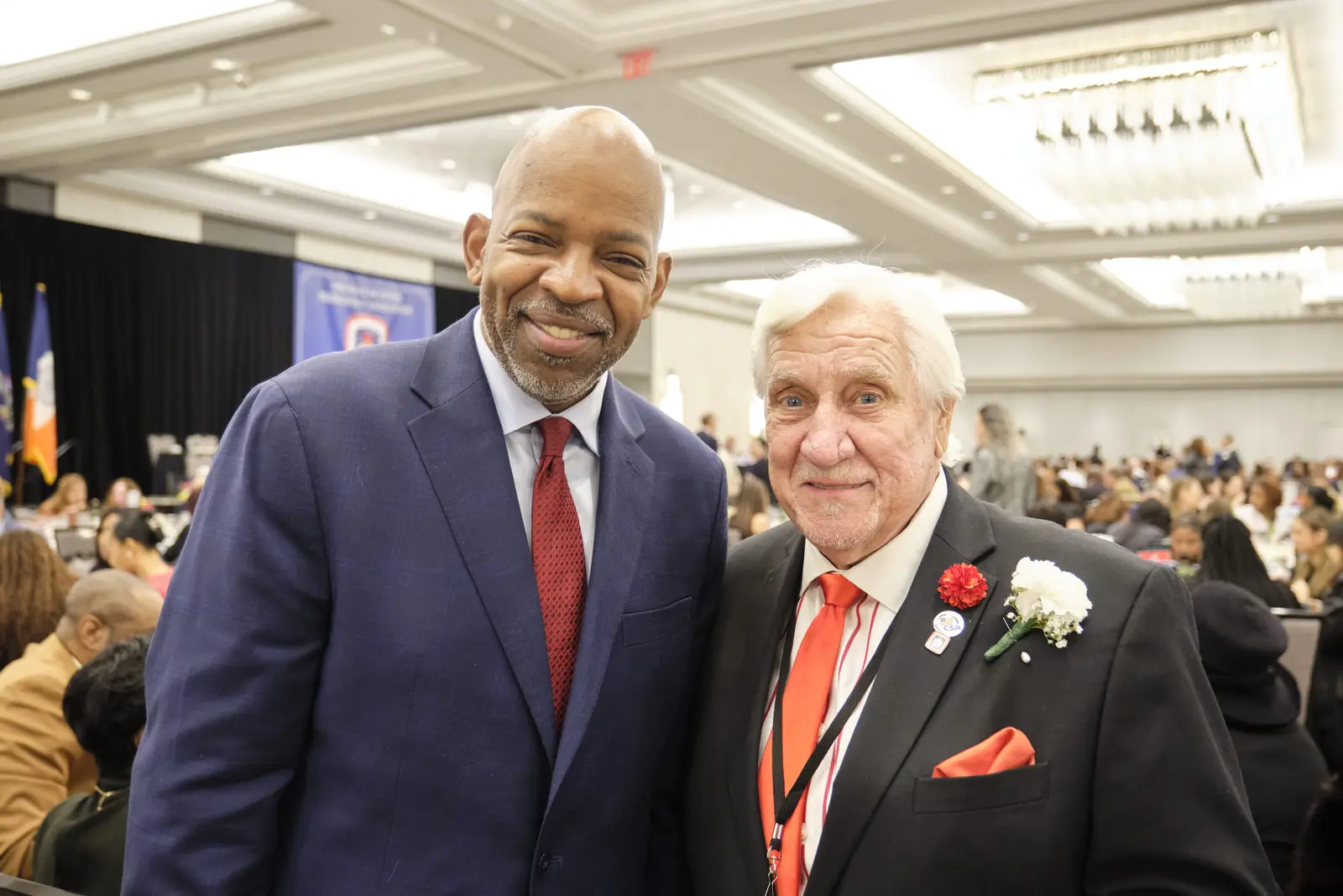 Two men in suits pose at formal event with attendees in background