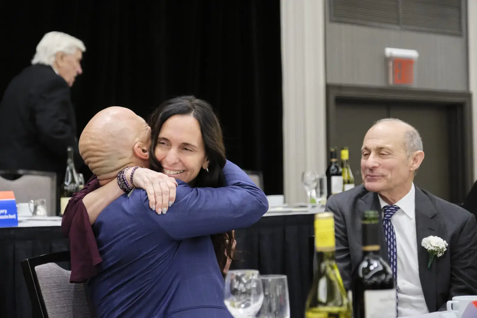 Two women hugging at formal event with guests seated nearby