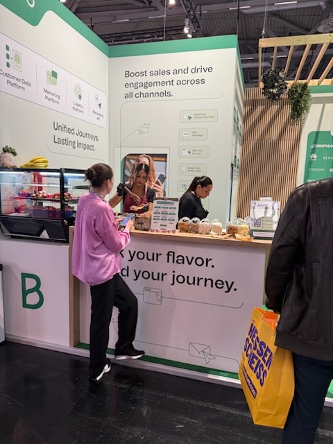 Messe-Stand von Brevo mit einem Tresen, an dem eine Food-Service-Mitarbeiterin Speisen serviert, und mehreren Besuchern vor dem Stand.