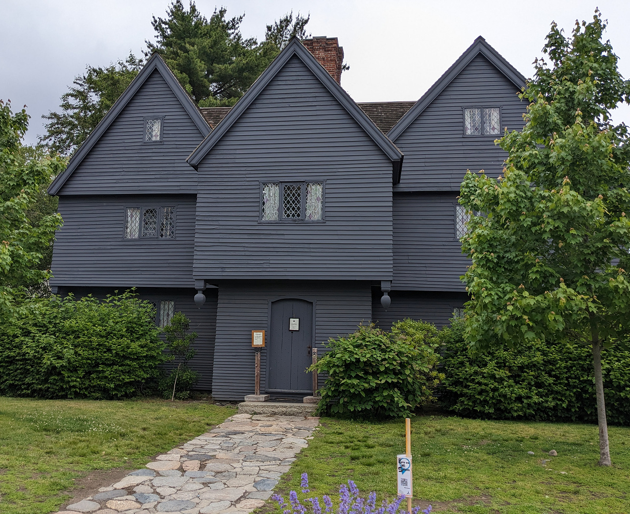 An old-looking black house with a stone path leading to the front door, surrounded by lush, green trees (The Witch House in Salem, Massachusetts)