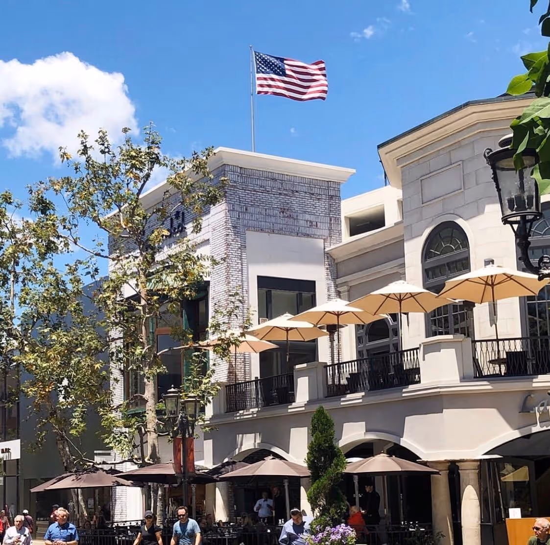 Outdoor shopping area with an American flag flying atop a brick building. White umbrellas and trees create a lively, sunny atmosphere, with people strolling below.