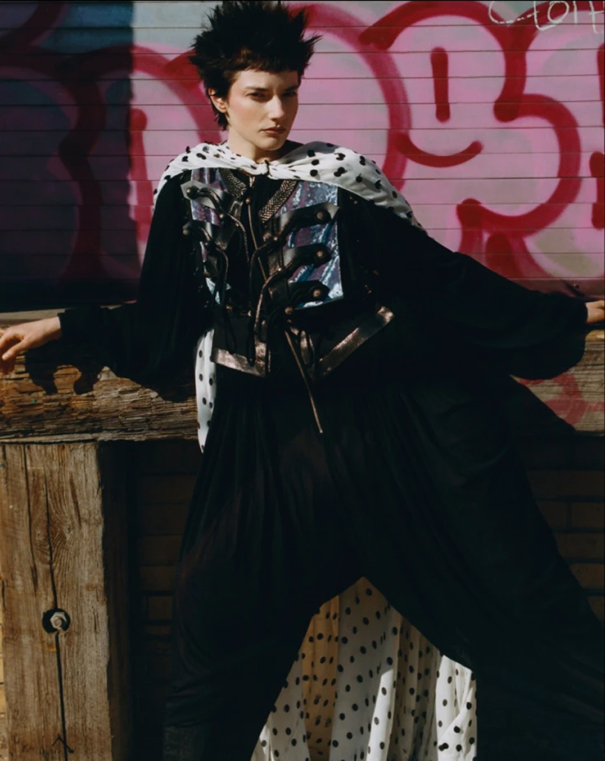 Person with spiky dark hair wearing a black outfit with a metallic vest and a white polka-dot cape standing against a rustic wooden wall with pink graffiti.