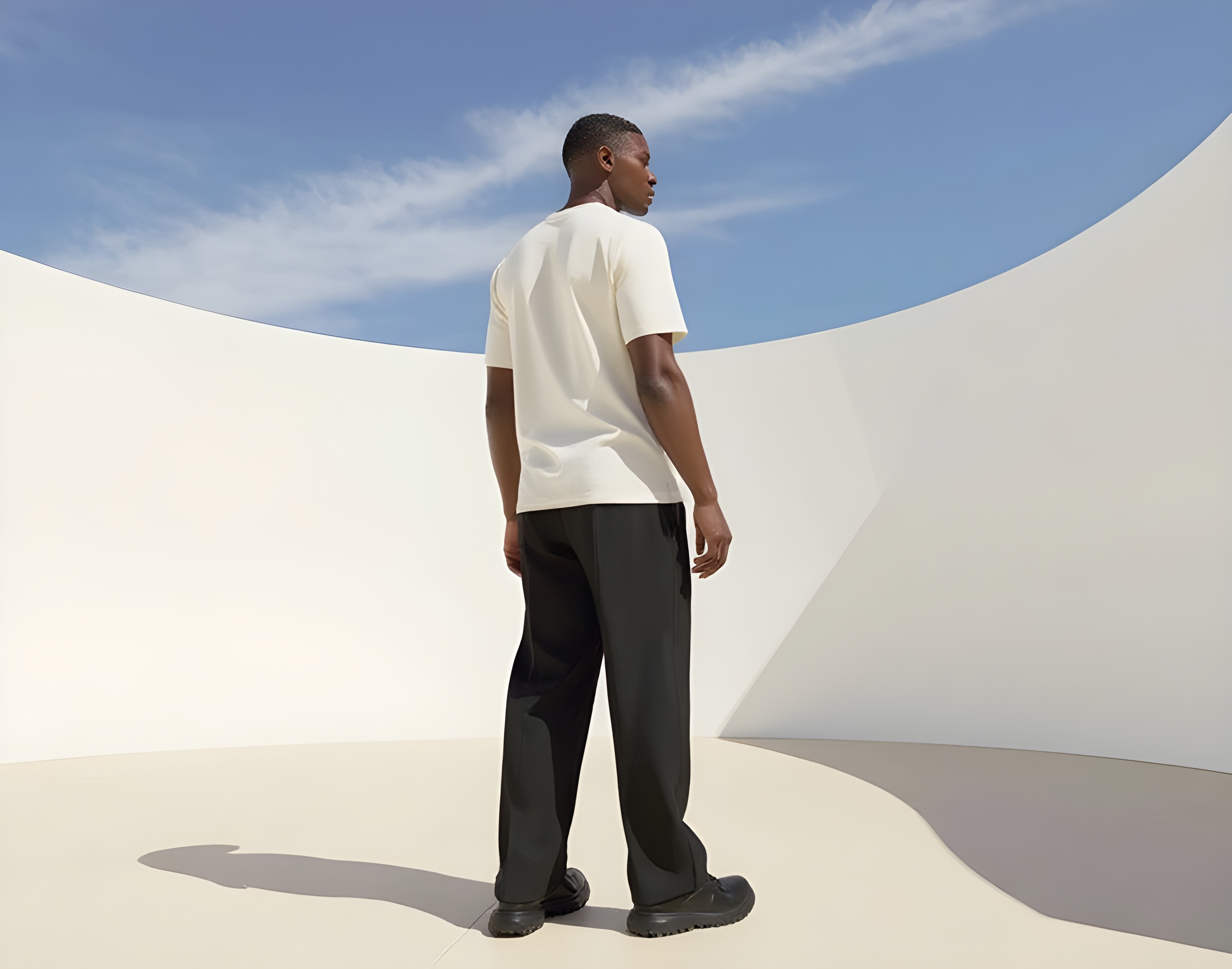 Man standing outdoors with back facing the camera, wearing a white t-shirt and black pants against a backdrop of curved white walls and blue sky.